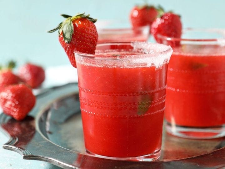 A silver tray holds three red drinks garnished with strawberries