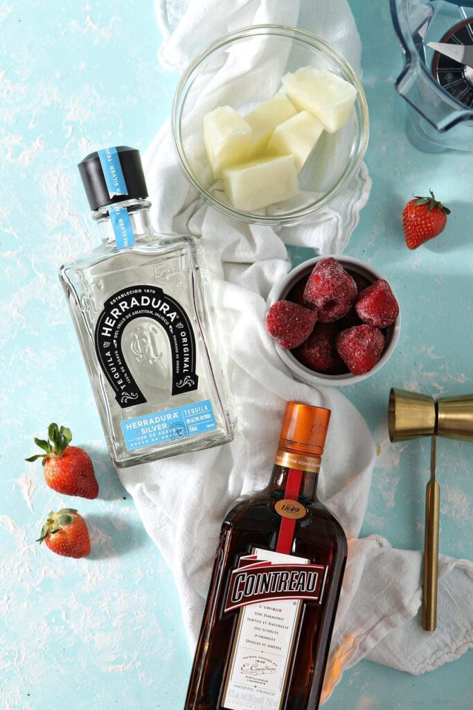 A bottle of tequila and a bottle of Cointreau sit on top of a white kitchen towel next to frozen strawberries and yellow ice cubes