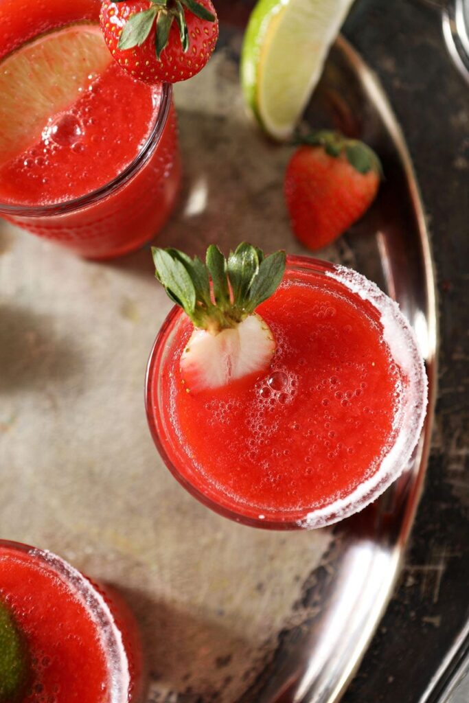 A halved strawberry floats on top of a frozen blended margarita recipe on a silver tray