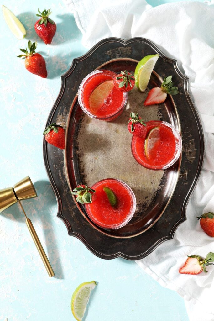 Three Frozen Strawberry Margaritas sit on a silver tray next to strawberries, lime wedges and a gold jigger