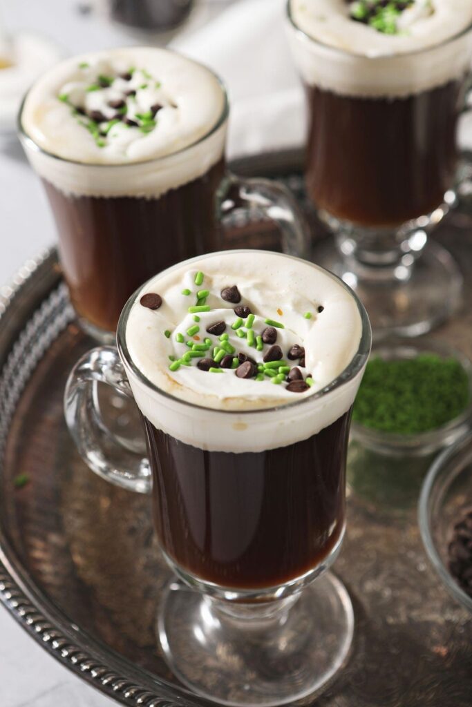 Three Irish Coffee drinks with whipped cream, chocolate and green sprinkles on a silver tray