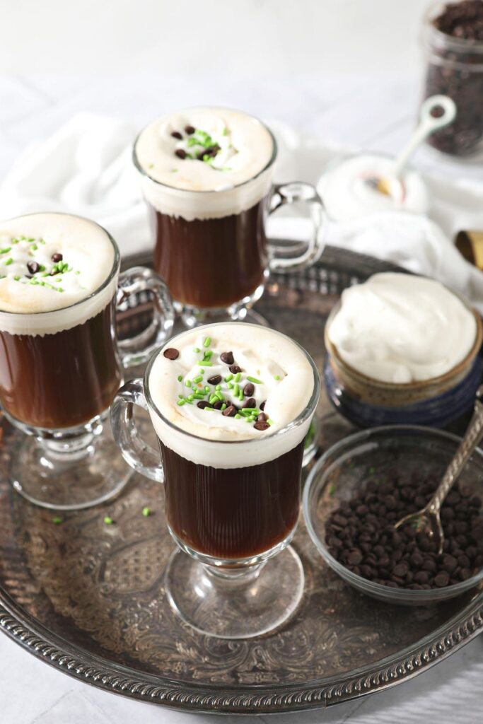 Three coffee drinks with whipped cream, chocolate and green sprinkles sit on a silver tray next to bowls of coffee beans and whipped cream