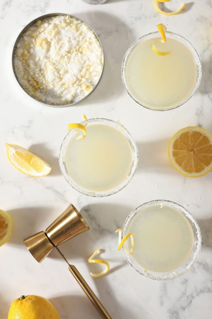 Three Lemon Drops sit on a marble surface next to lemon sugar, lemon wedges and a golden jigger