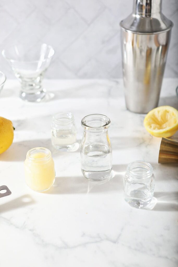 Lemon juice, simple syrup, vodka and Cointreau in bottles on a marble surface surrounded by other mixed drink materials