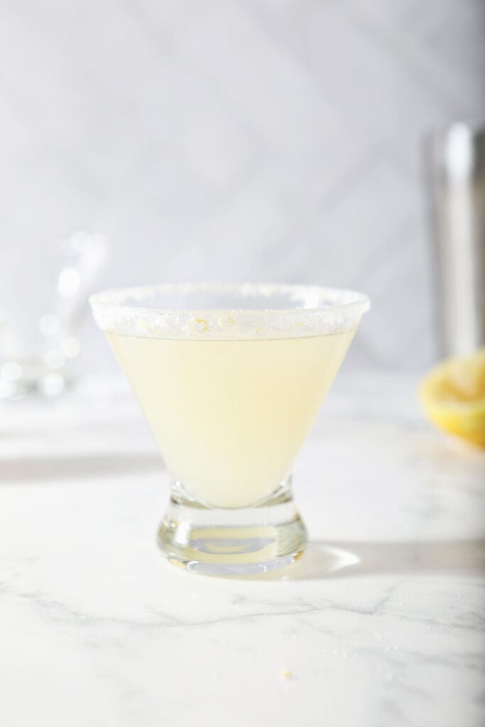 A single Lemon Drop Martini sits on a marble surface in front of a cocktail shaker and a lemon wedge