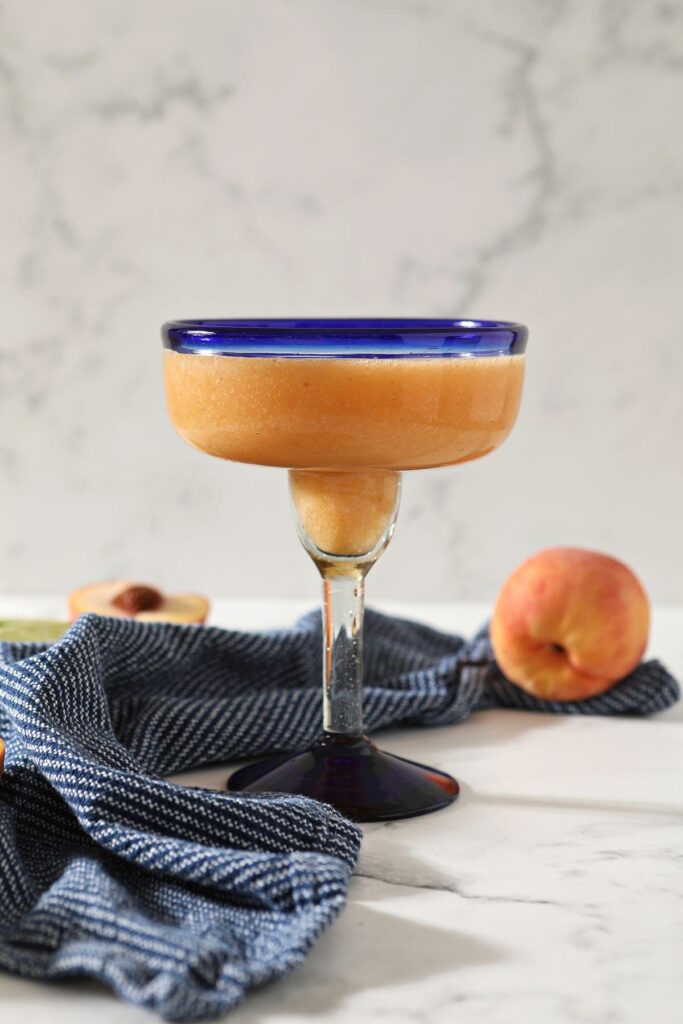 Homemade peach margaritas are a wonderful summertime sipper! This 5-ingredient peach margarita recipe involves real peaches (not peach-flavored liqueur) and mixes up in a blender in less than 10 minutes. Makes 2 drinks, but is easily scalable.   A peach margarita in a blue-rimmed margarita glass