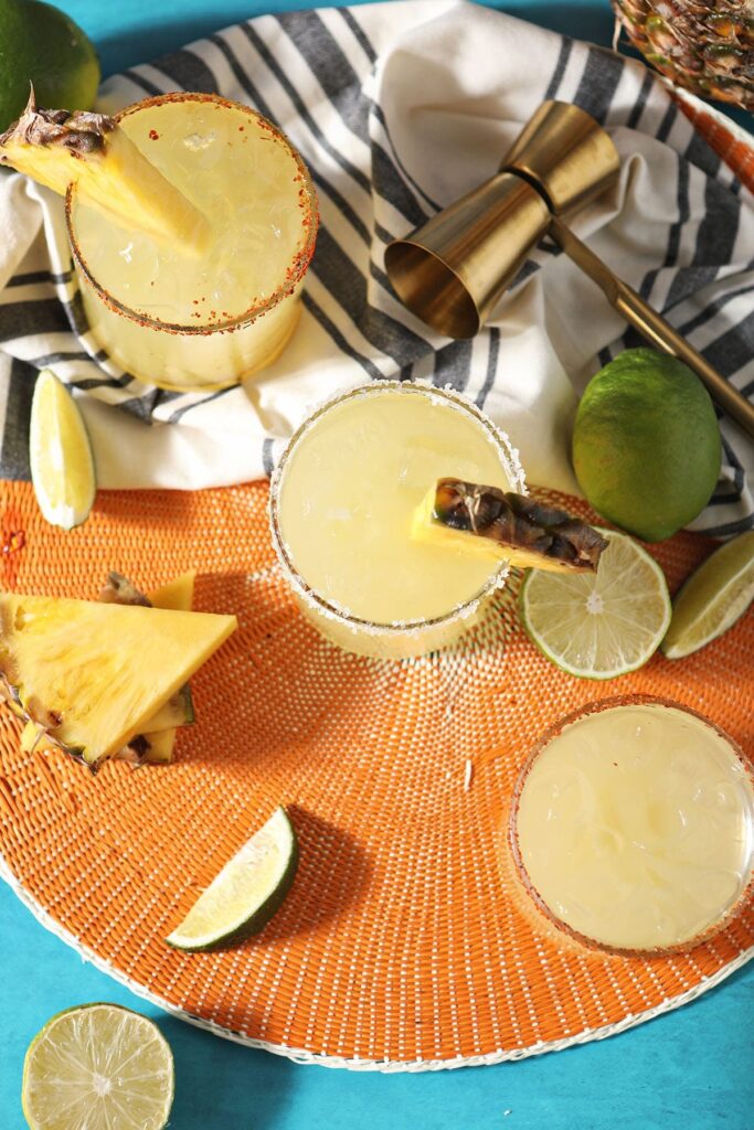 Three pineapple drinks with lime and pineapple wedges on an orange mat