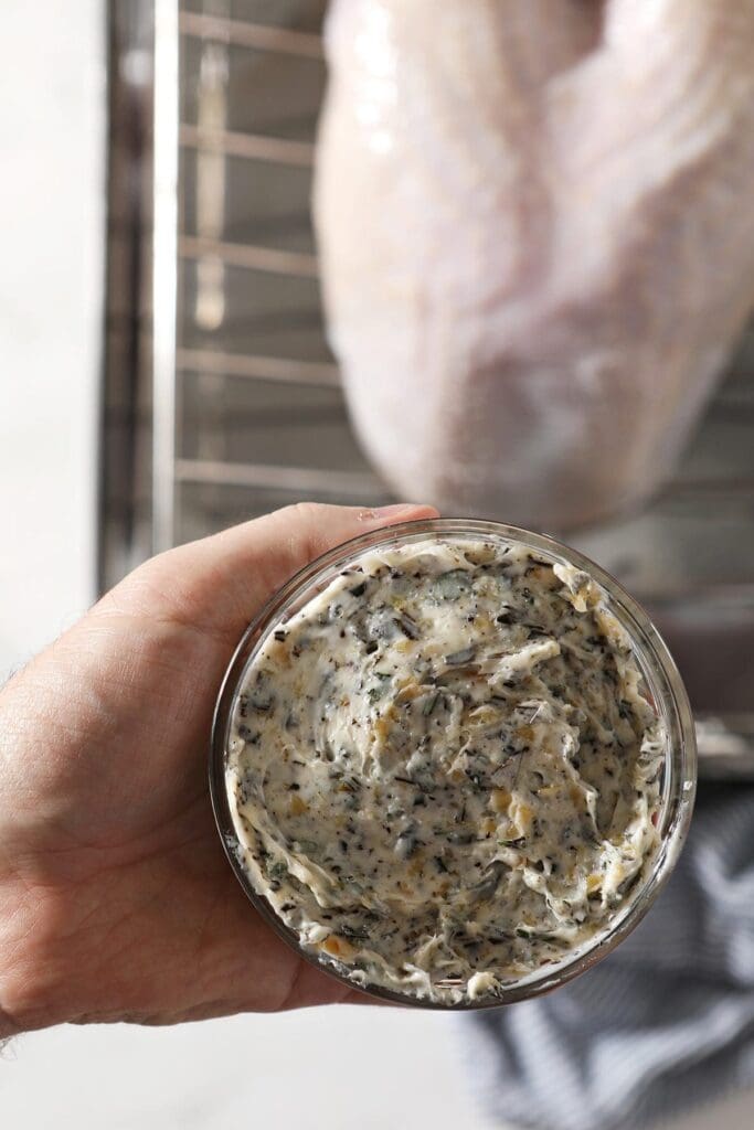 A hand holds a container of herb butter to rub on a turkey breast above the turkey