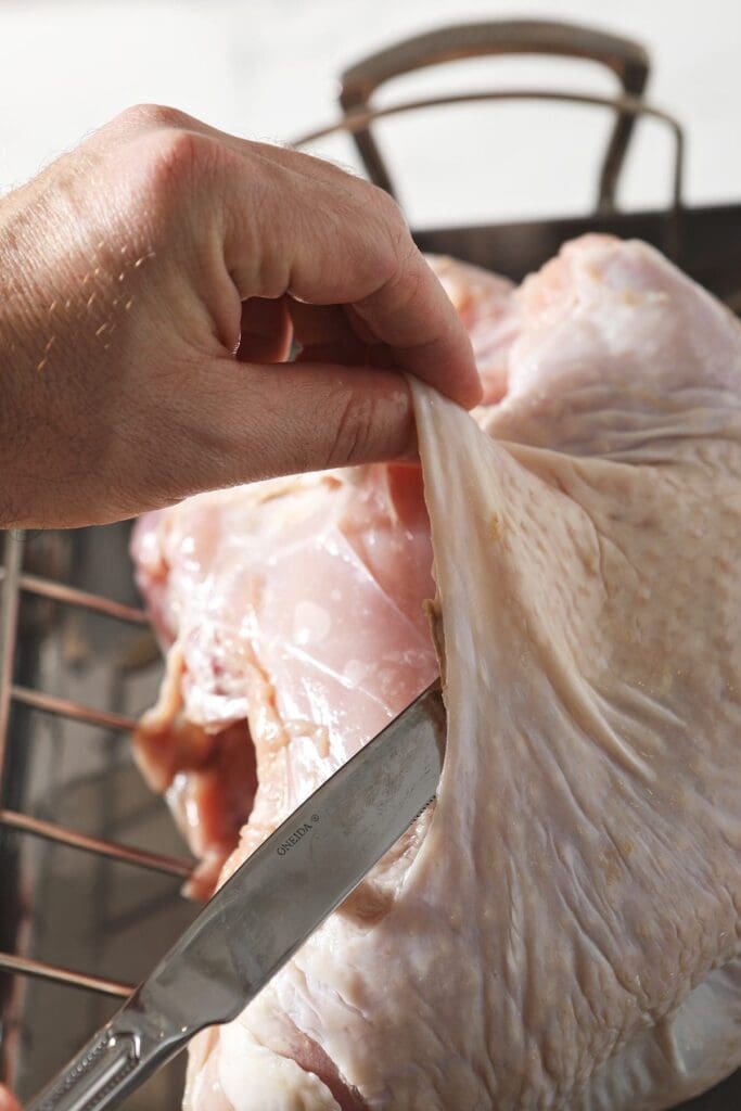 A hand uses a butter knife to pull the turkey skin away from the meat to add butter to