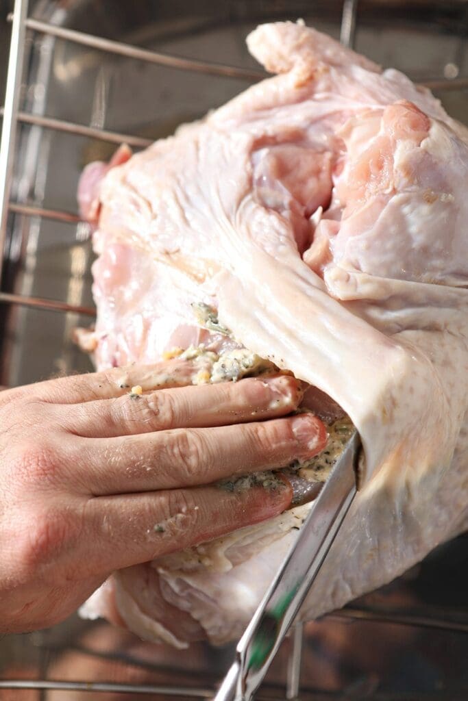 A hand rubs herb butter onto a raw turkey breast on a roasting pan