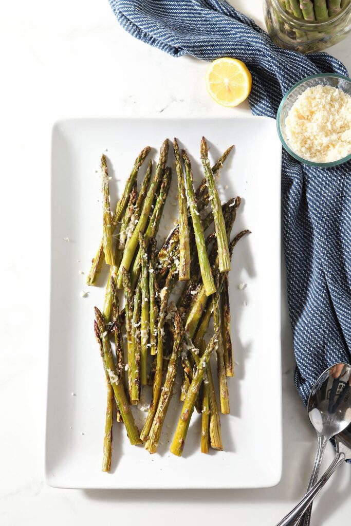 Spears of broiled asparagus on a white platter next to a bowl of parmesan and lemon