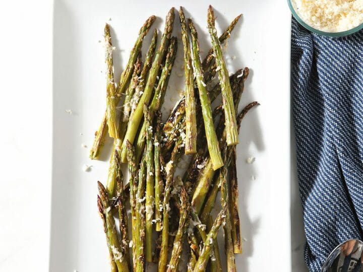 Spears of broiled asparagus on a white platter next to a bowl of parmesan and lemon