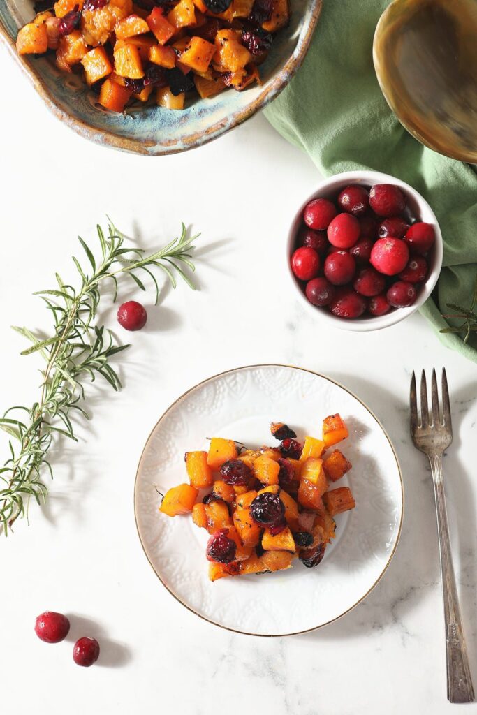 A plate of roasted butternut squash with a fork near cranberries and rosemary