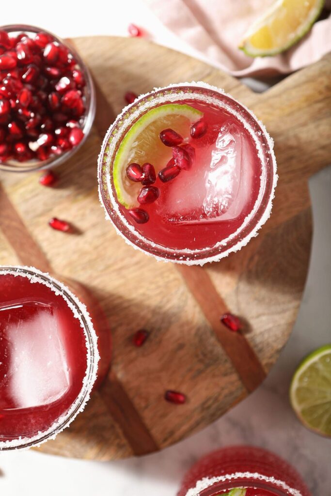 Two pomegranate margaritas on wood garnished with lime and pomegranate arils