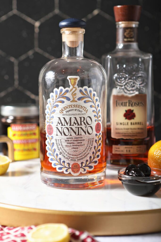 Bottles of amaro and bourbon with luxardo cherries and a lemon