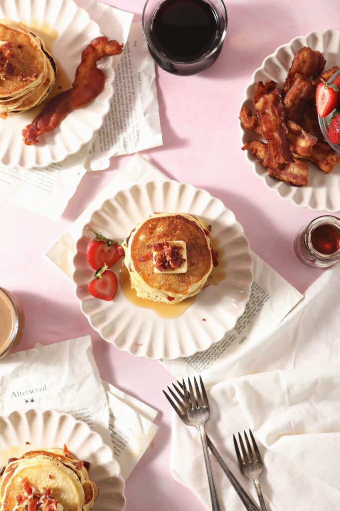 Three plates of homemade pancakes with bacon on white plates and a pink countertop