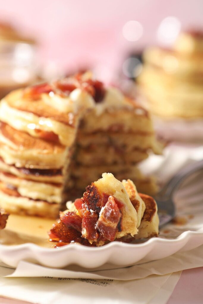 A fork holds a bite of bacon pancakes next to a stack of pancakes