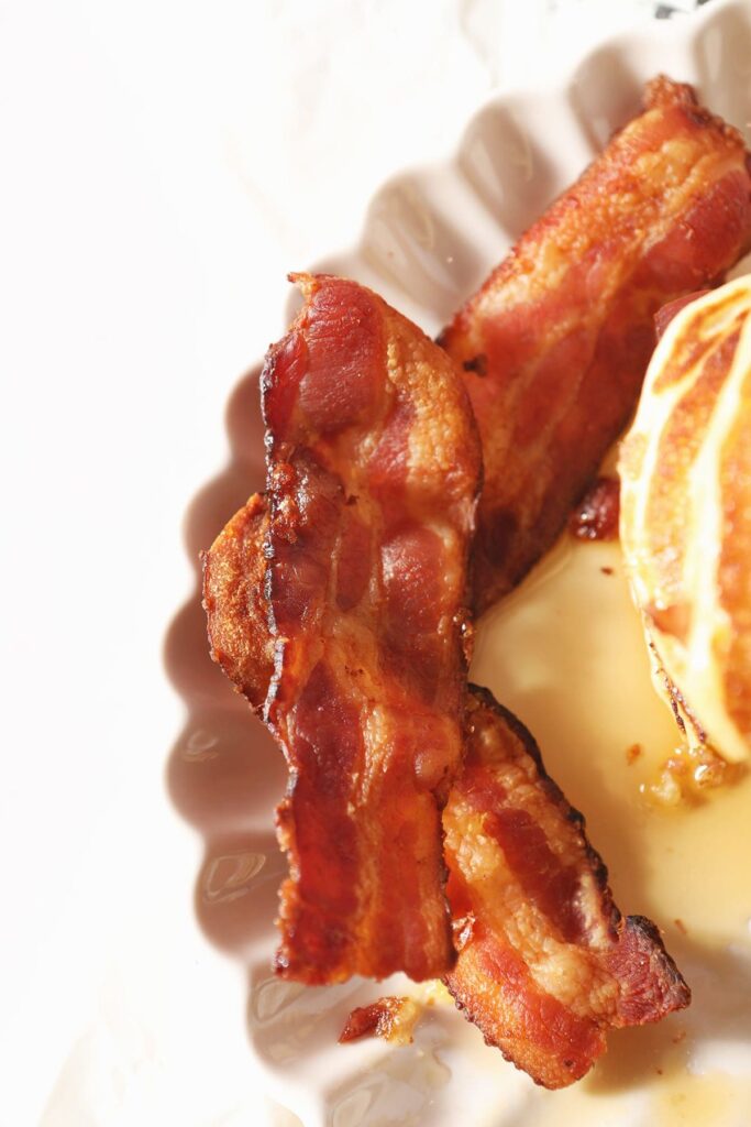 Close up of cooked bacon on a plate next to pancakes and syrup