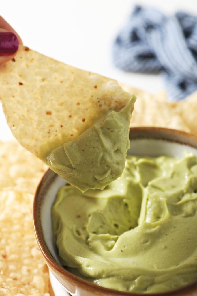 A tortilla chip dips into a bowl of avocado cream