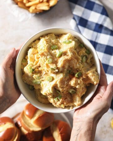 A woman holds a bowl of traditional potato salad over a table of other BBQ dishes