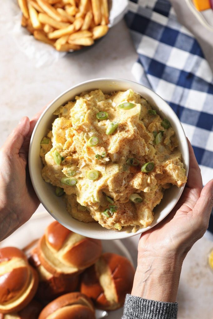 A woman holds a bowl of traditional potato salad over a table of other BBQ dishes