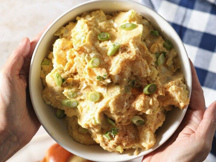 A woman holds a bowl of traditional potato salad over a table of other BBQ dishes