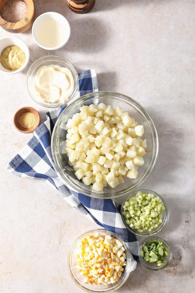 Potato salad ingredients in bowls on a countertop with a blue checked towel