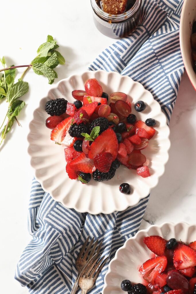 Overhead of a white plate holding a serving of Summer Fruit Salad next to mint, forks and a geometric blue towel