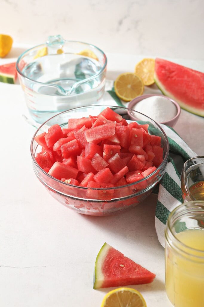 A bowl of cubed watermelon with other lemonade ingredients on marble