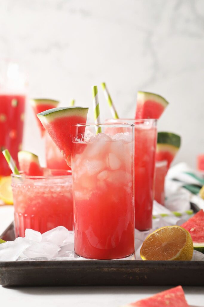 Four glasses of watermelon lemonade on a tray with ice and lemons and watermelon wedges