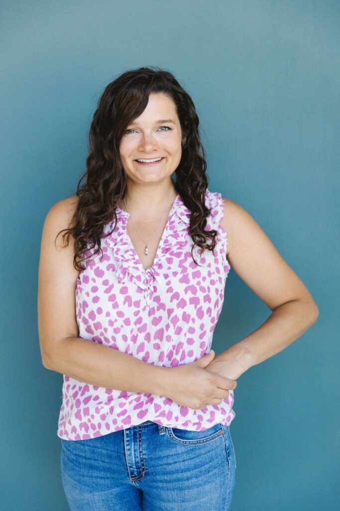A woman with dark curly hair wearing a purple and white polka dot top stands in front of a turquoise wall