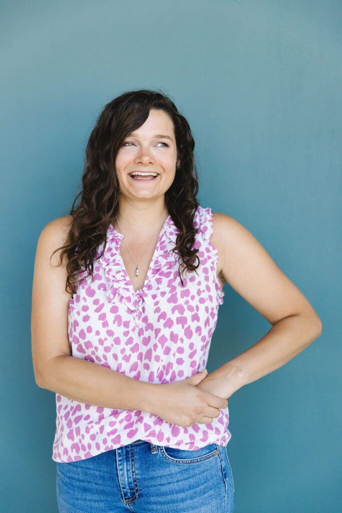 A woman with dark curly hair wearing a purple and white polka dot top laughs and looks off in the distance as she stands in front of a turquoise wall