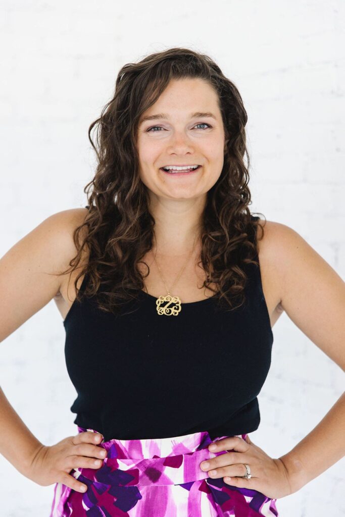 A woman with dark curly hair wearing a black tank top in front of a white wall