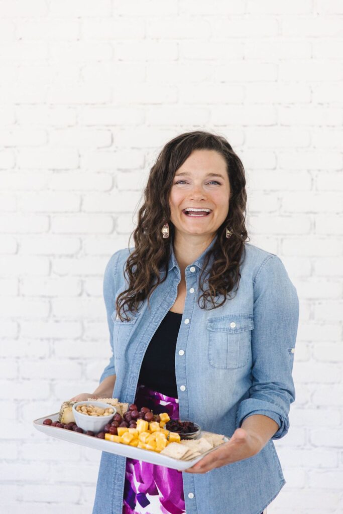 A dark-haired woman wearing a jean shirt over a black tank holds a cheeseboard and laughs by a table