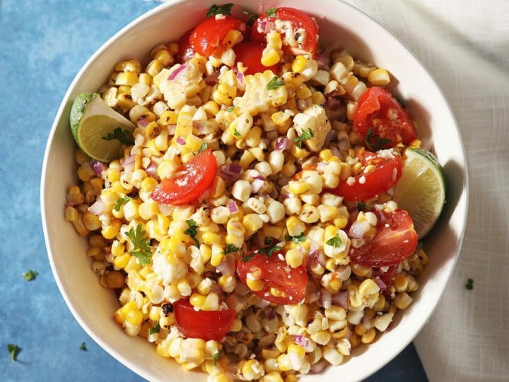 A bowl of corn salsa with tomatoes and limes on a blue countertop