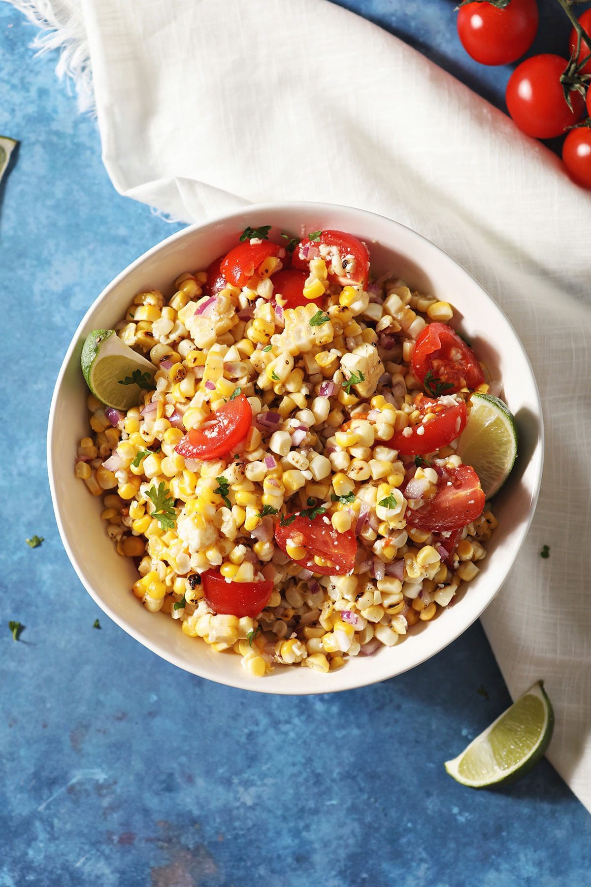 A bowl of corn salsa with tomatoes and limes on a blue countertop