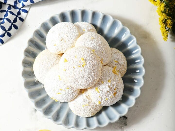 A blue plate of Lemon Cooler Cookies next to flowers and lemon slices