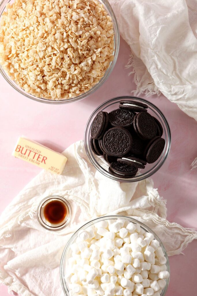 Oreos and other crispy rice treat ingredients in bowls on a pink surface