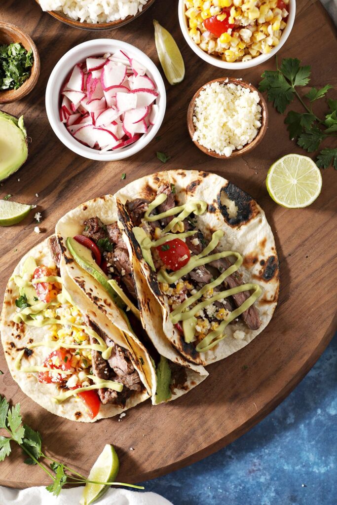 Three skirt steak tacos surrounded by toppings in bowls on a wooden board