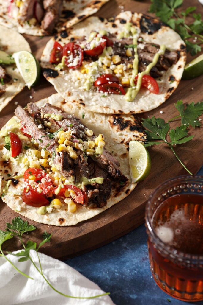 Three skirt steak tacos on a wooden board next to a cup of beer