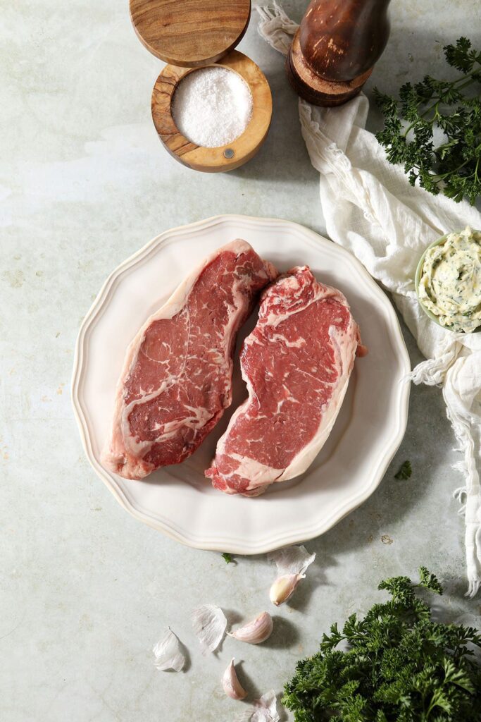 Two steaks on a white plate surrounded by herbs, garlic butter, salt and pepper