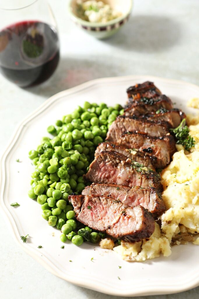 Sliced steak on a white plate with peas and mashed potatoes