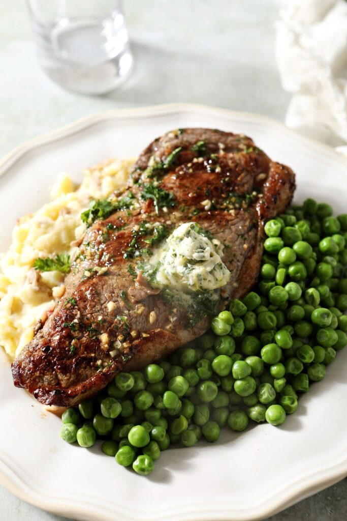 A whole steak topped with a scoop of herb butter for steak on a plate with peas and mashed potatoes