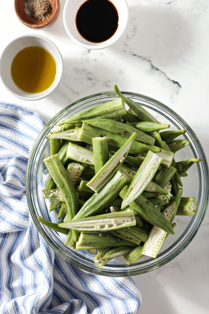 Okra in a bowl with a blue striped towel and other ingredients in bowls