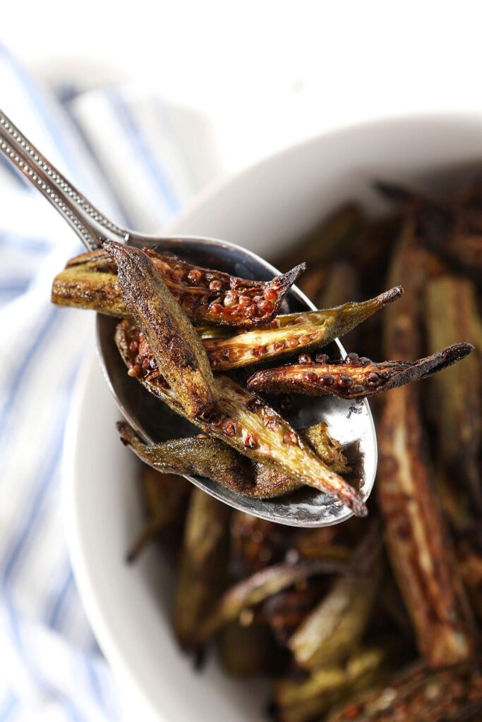 A spoon holds a serving of roast okra above a bowl and a blue striped towel