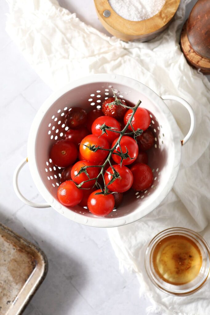 A white colander holds vine-ripened cherry tomatoes surrounded by ingredients in bowls