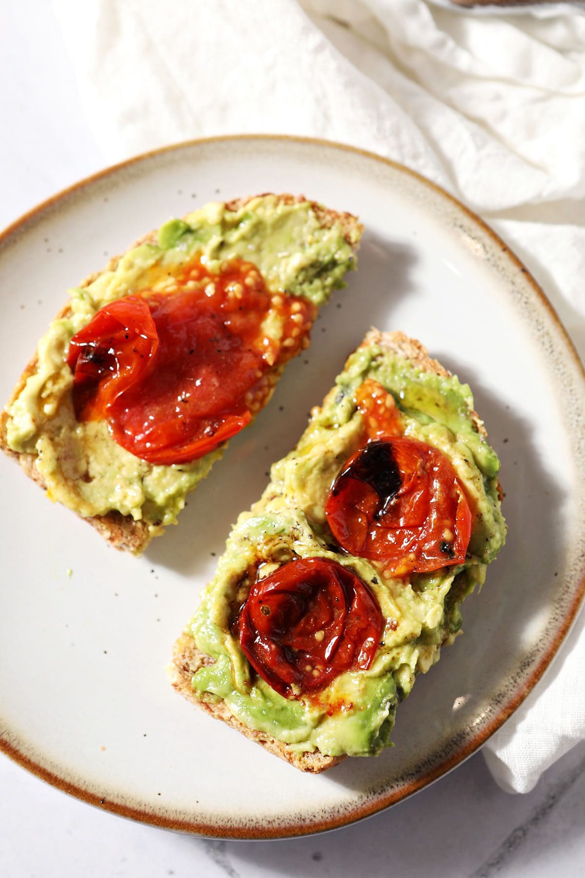 These Oven Roasted Tomatoes are the perfect way to enjoy the sweetness of summer. All you need are four ingredients and 20 minutes to make the most decadent roasted cherry tomatoes around! Add them to a sandwich or serve them alongside a meal to make it extra special. Avocado toast sliced in half with roasted tomatoes on top on a plate