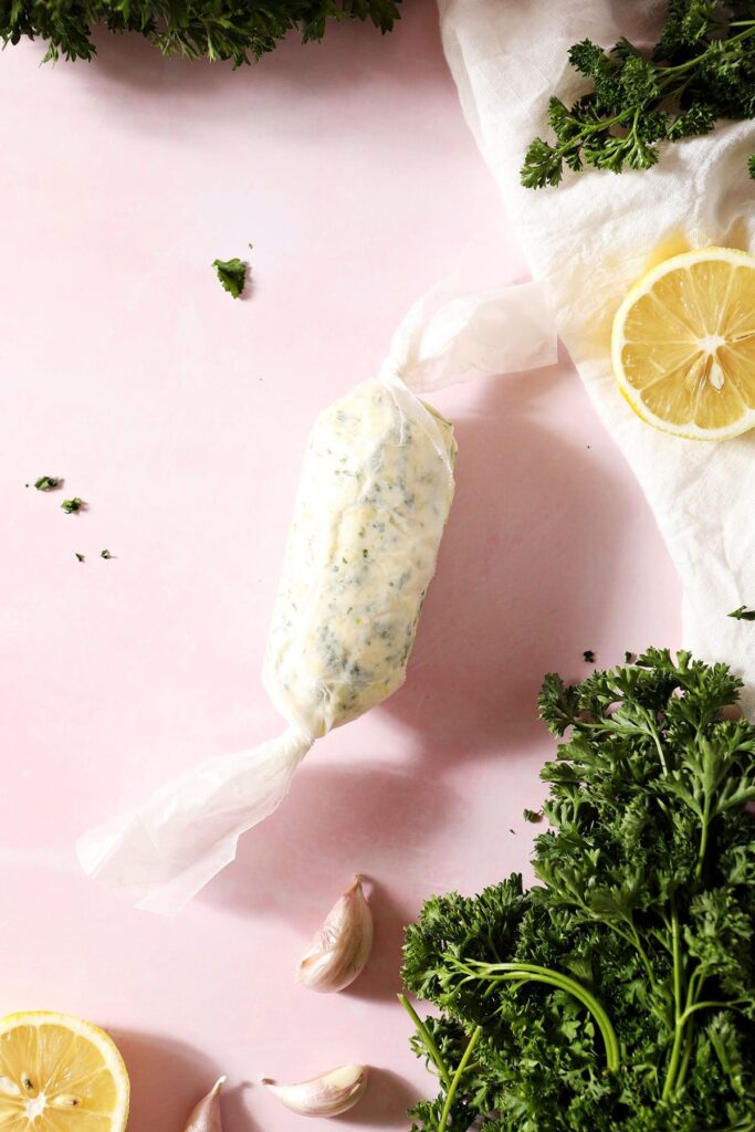 Garlic Herb Compound Butter wrapped in wax paper on a pink countertop surrounded by herbs, garlic and lemon