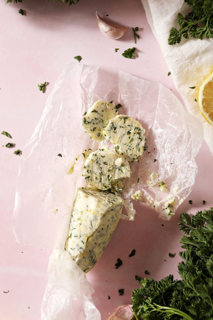 A log of garlic herb butter and slices of it on wax paper surrounded by herbs, lemons and garlic