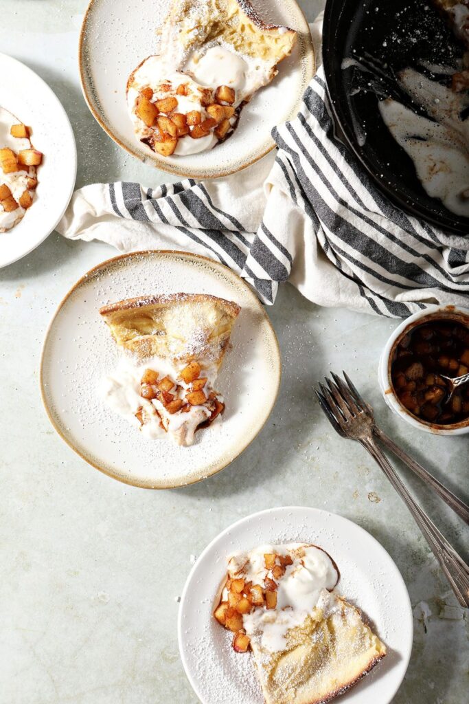 Slices of a German pancake on plates next to a skillet holding it, garnished with whipped cream, apples and powdered sugar